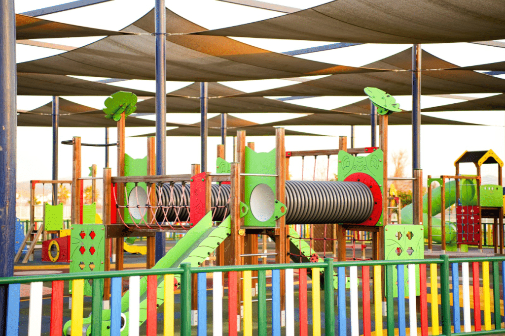 Parque infantil con estructuras multijuego, toboganes y túneles, protegido por toldos tensados que proporcionan sombra sobre las áreas de juego.