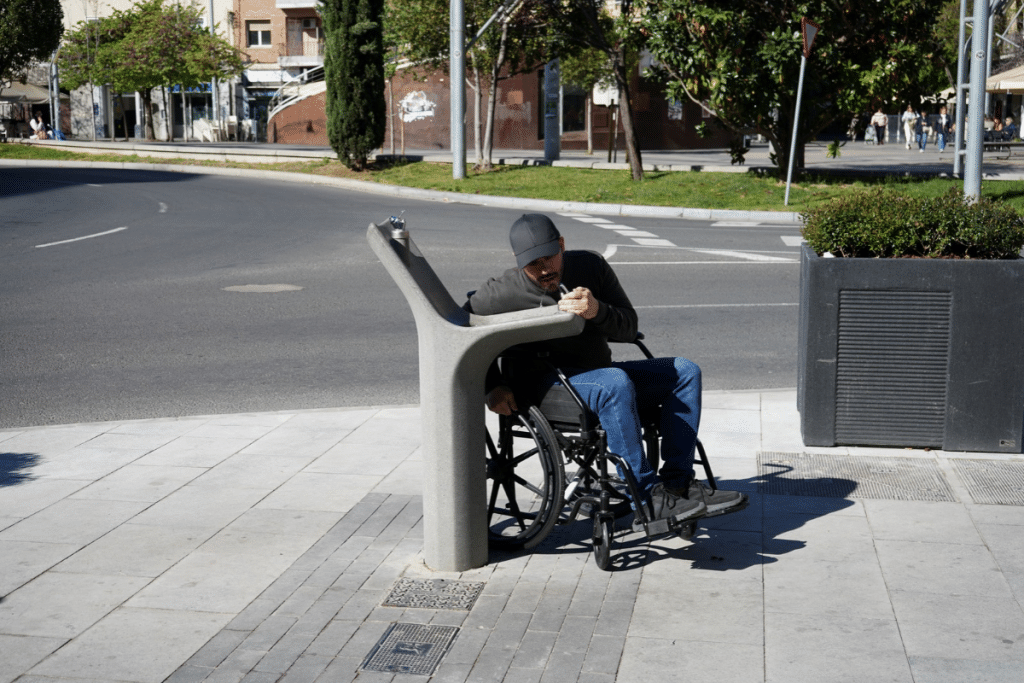 Hombre en silla de ruedas utilizando una fuente de agua accesible instalada en un espacio público urbano.