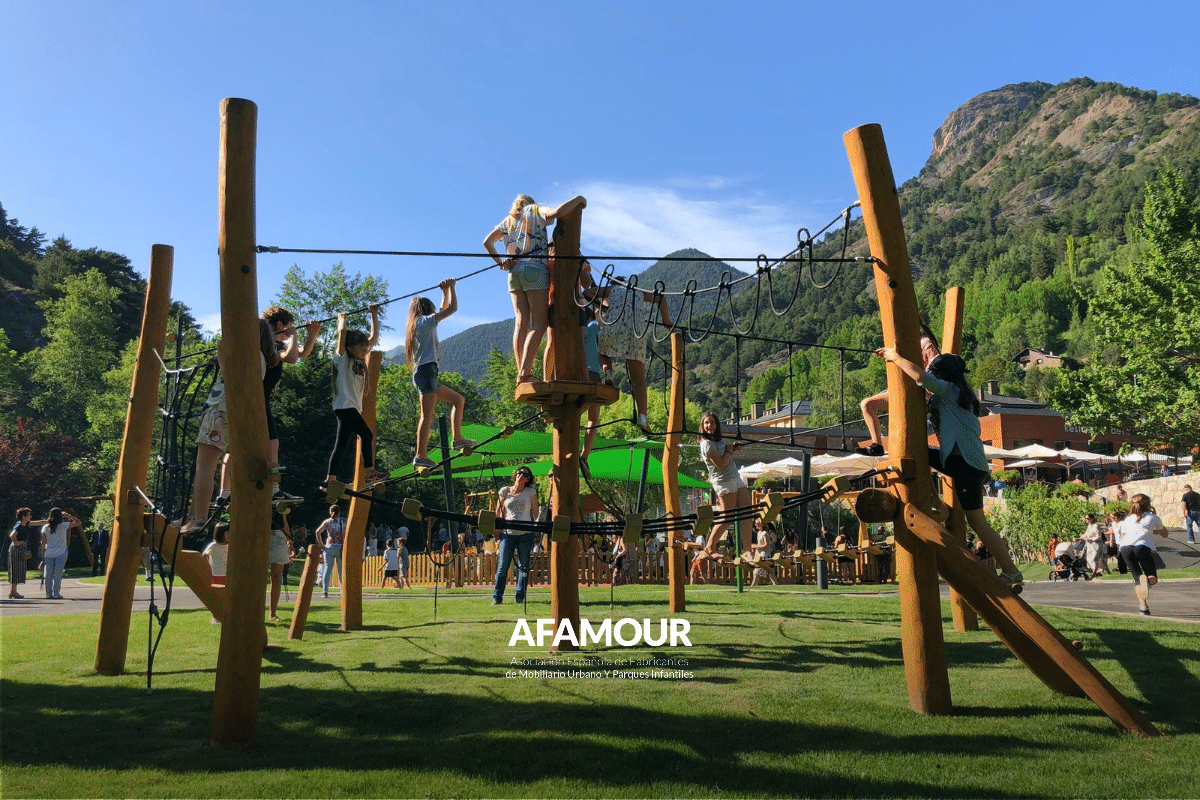 Niños jugando en una estructura de cuerdas y madera en un parque infantil al aire libre, con césped, árboles y montañas al fondo.